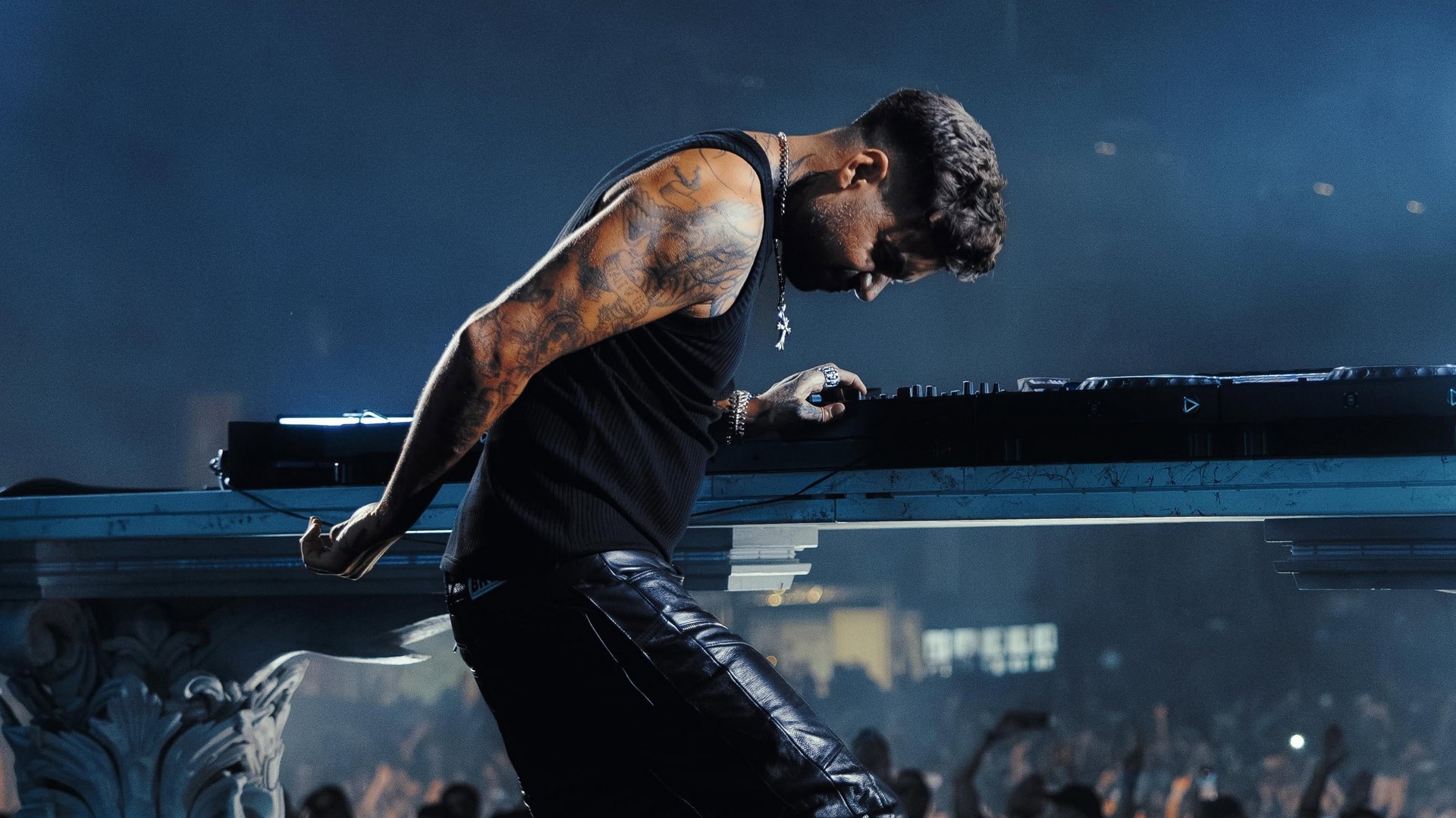 Dj anyma aux bras tatoués mixe sur une console devant une foule dans une salle de concert sombre