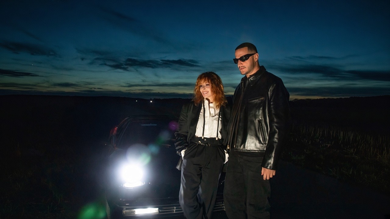 Mylène Farmer et DJ Snake habillées en noir posent de nuit devant une voiture aux phares allumés